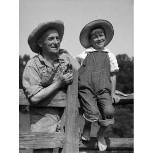  on Fence With Man Standing Beside Him, Both Wearing Bib Overalls 